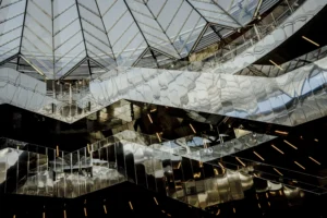 Cette photographie met en scène une structure architecturale moderne, probablement un atrium ou un hall couvert. Le sujet principal est l’intersection des poutres métalliques et des panneaux de verre formant une géométrie complexe.La composition joue sur les lignes diagonales et les jeux d’ombres. Le cadrage serré accentue la profondeur et la symétrie. La lumière, tamisée et artificielle, crée des contrastes nets entre ombres et reflets. Les couleurs dominantes sont les gris métalliques, les blancs et les teintes bleutées, renforçant une ambiance industrielle et futuriste. Le style est épuré et minimaliste, évoquant une atmosphère à la fois froide et élégante.