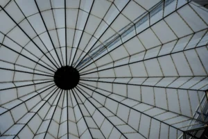 Cette photographie montre une structure géométrique en forme de dôme ou de parasol, vue de dessous. Le sujet principal est le motif radial formé par les barres métalliques convergeant vers un point central noir.La composition est symétrique et dynamique, avec un cadrage serré qui accentue les lignes convergentes. La lumière, naturelle et diffuse, crée des contrastes doux entre ombres et clarté. Les couleurs dominantes sont des tons gris et blancs, renforçant une ambiance minimaliste et industrielle.Le style est épuré, presque abstrait, évoquant une sensation de profondeur et d'ordre architectural. L'ambiance est à la fois moderne et contemplative.