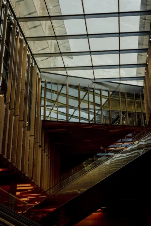 Cette photographie met en scène un **escalier mécanique** dans un espace couvert, probablement une gare ou un centre commercial. La **composition** est dynamique, avec des lignes convergentes menant vers le haut.Le **cadrage** est serré, capturant une perspective verticale et légèrement inclinée. La **lumière**, naturelle et tamisée, filtre à travers une **verrière**, créant des jeux d'ombres et de transparence. Les **couleurs dominantes** sont des tons froids (gris, bleu pâle) et des reflets métalliques.Le style est **minimaliste et industriel**, évoquant une ambiance **moderne et fonctionnelle**, presque mélancolique.