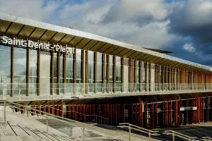 Cette photographie montre la gare **Saint-Denis - Pleyel**, sujet principal de l'image. La composition met en valeur la structure moderne avec une façade en verre et métal, soulignée par une grande toiture en bois. Le cadrage est large, incluant une partie du ciel nuageux.La lumière naturelle éclaire la scène, créant des contrastes entre ombres et reflets. Les couleurs dominantes sont les tons gris (métal et béton), bruns (bois) et quelques touches de rouge. Le style architectural est contemporain et fonctionnel. L’ambiance est dynamique et urbaine, reflétant une infrastructure moderne et bien intégrée.