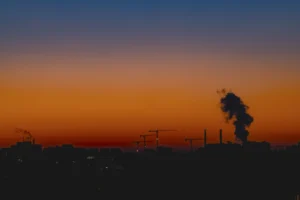 Cette photographie capture un coucher de soleil dramatique. Le **sujet principal** est l’énorme panache de fumée noire s’élevant à l’horizon, contrastant avec le ciel. La **composition** est épurée, avec une ligne d’horizon basse et des éléments urbains indistincts en silhouette.Le **cadrage** est large, mettant en valeur l’immensité du ciel. La **lumière** est tamisée, avec des reflets dorés et orangés. Les **couleurs dominantes** sont les tons chauds du crépuscule (orange, rouge) et les bleus profonds du ciel.Le **style** est réaliste et documentaire, évoquant une **ambiance** à la fois apocalyptique et mélancolique, mêlant beauté naturelle et menace industrielle.