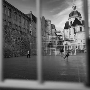 Pascal SC · 01/11/2025 · 1920 x 1920 Cette photographie en noir et blanc capture une scène urbaine dynamique. Le sujet principal est un enfant jouant au ballon dans une cour, entouré d’un mur en pierre et d’un bâtiment historique avec une flèche distinctive en arrière-plan.La composition utilise un cadrage serré, encadré par une fenêtre ou un objet rectangulaire en bas. La lumière est douce, créant des contrastes marqués entre ombres et clartés. Les tons dominants sont les gris et noirs, renforçant une ambiance nostalgique et intemporelle.Le style est documentaire et spontané, évoquant une atmosphère à la fois simple et poétique.