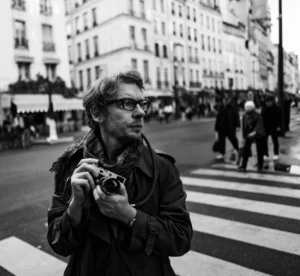 Pascal SC · 01/11/2025 · 1920 x 1767 Cette photographie en noir et blanc met en scène un homme comme sujet principal, vêtu d’un manteau et tenant un appareil photo. La composition est centrée sur lui, avec un cadrage serré qui isole le sujet des éléments arrière-plan.L’ambiance est urbaine, avec des bâtiments et des passants flous en arrière-plan. La lumière semble naturelle, probablement en journée, créant des contrastes doux. Le style est documentaire et intime, évoquant une atmosphère authentique et spontanée. Les tons dominants sont les gris et noirs, renforçant l’aspect classique et intemporel de l’image.