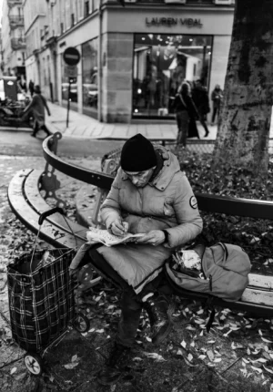 Pascal SC · 01/11/2025 · 1340 x 1920 Cette photographie en noir et blanc met en scène un homme assis sur un bord de trottoir, penché sur un document. Il semble être un militaire ou un ancien combattant, reconnaissable à son uniforme.La composition est centrée sur lui, avec des éléments urbains en arrière-plan (boutiques, passants). Le cadrage est serré, mettant l’accent sur son activité et son environnement immédiat. La lumière est naturelle, contrastée, accentuant les ombres et les détails.Les couleurs dominantes sont les nuances de gris, typiques du noir et blanc, renforçant une ambiance mélancolique et introspective. Le style est documentaire, réaliste, évoquant une scène de vie quotidienne.