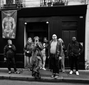 Pascal SC · 01/11/2025 · 1920 x 1839 Cette photographie en noir et blanc montre un groupe de cinq personnes posant devant une porte d’immeuble. Le sujet principal est leur attitude décontractée et dynamique, avec des poses théâtrales.La composition est symétrique, avec les individus alignés. Le cadrage est serré, mettant l’accent sur eux et l’entrée sombre derrière eux. La lumière est naturelle, contrastant les visages éclairés et l’arrière-plan ombré.Les couleurs dominantes sont les nuances de gris, renforçant une ambiance rétro et cinématographique. Le style évoque un mélange de street style et de pose artistique, créant une ambiance à la fois décontractée et légèrement provocante.