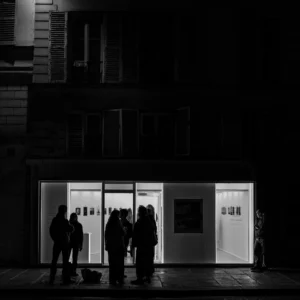 Cette photographie en noir et blanc montre un groupe de personnes devant une galerie d'art ou une exposition nocturne. Le sujet principal est l'interaction des visiteurs devant les œuvres éclairées.La composition repose sur un cadrage serré, mettant en valeur les silhouettes et les vitrines lumineuses. La lumière, contrastée, éclaire uniquement l’espace d’exposition, créant une ambiance mystérieuse et dramatique. Les couleurs dominantes sont les tons de gris, renforçant l’atmosphère nocturne et artistique. Le style est épuré et moderne, évoquant une ambiance contemplative et élégante.