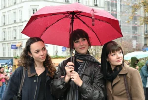 Mary · 15/11/2025 · 2784 x 1881 Cette photographie montre trois jeunes femmes comme sujet principal, souriantes et regroupées sous un parapluie rouge. La composition est symétrique, avec les femmes alignées au centre. Le cadrage est serré, mettant l’accent sur elles.La lumière semble naturelle, probablement en journée. Les couleurs dominantes sont les tons neutres des vêtements et le rouge vif du parapluie. L’ambiance est joyeuse et conviviale, renforcée par leurs expressions souriantes et les couleurs vives. Le style est spontané et photographique, typique d’un moment de vie quotidienne.