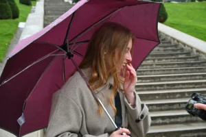 Mary · 15/11/2025 · 2784 x 1856 Cette photographie met en scène une jeune femme comme sujet principal, tenant un parapluie marron foncé. Elle semble parler au téléphone, le regard dirigé vers le bas.La composition est simple et centrée, avec un cadrage serré sur le visage et le haut du corps. L’arrière-plan montre des escaliers et un environnement extérieur légèrement flou.La lumière est naturelle, probablement en journée, avec des tons doux. Les couleurs dominantes sont les teintes neutres (gris du manteau) et le marron du parapluie, adoucies par des touches de vert dans l’arrière-plan.L’ambiance est calme et intime, évoquant une scène du quotidien.