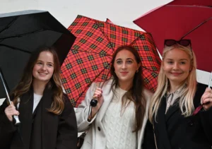 Mary · 15/11/2025 · 2784 x 1957 Cette photographie montre trois jeunes femmes souriantes tenant des parapluies. Le sujet principal est leur joie et leur interaction.La composition est symétrique, avec les trois femmes alignées côte à côte. Le cadrage est serré, mettant en valeur leurs visages et leurs expressions. La lumière semble naturelle et douce, éclairant uniformément les sujets.Les couleurs dominantes sont les tons froids (noir, gris) et chauds (rouge plaid, blanc). L’ambiance est joyeuse et décontractée, évoquant une scène conviviale et légère. Le style est simple et spontané.