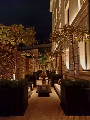 Cette photographie montre une élégante cour intérieure nocturne. Le sujet principal est l’allée bordée de haies et de plantes grimpantes éclairées, menant vers un bâtiment classique. La composition est symétrique, avec des colonnes et des balcons ornés.Le cadrage est serré, guidant le regard vers le fond. La lumière douce et chaude, provenant des lanternes et des éclairages discrets, crée une ambiance chaleureuse et sophistiquée. Les couleurs dominantes sont les tons verts des plantes, les ors des ornements et les teintes beige et crème des murs, renforçant un style classique et luxueux. L’ambiance est à la fois intimiste et raffinée.