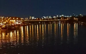 Cette photographie capture un **pont illuminé** enjambant une rivière la nuit. Le **sujet principal** est le pont, dont les **lumières chaudes** créent des reflets dorés sur l’eau.La **composition** est équilibrée, avec une perspective symétrique et des lignes directrices menant vers l’horizon. Le **cadrage** est large, incluant les bâtiments éclairés en arrière-plan.Les **couleurs dominantes** sont des tons **jaunes, oranges et bleus nocturnes**. Le style est **réaliste et photographique**, avec une ambiance **sereine et légèrement magique**, évoquant une ville paisible sous les lumières artificielles.