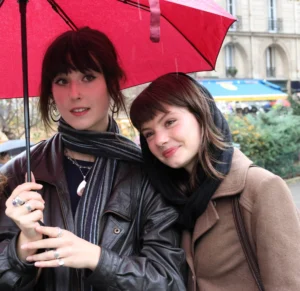 Laurence · 4786 x 4640 Cette photographie montre deux jeunes femmes souriantes sous un parapluie rouge. Le sujet principal est leur complicité et leur proximité.La composition est symétrique, avec un cadrage serré sur elles. La lumière semble naturelle, légèrement tamisée, créant des ombres douces. Les couleurs dominantes sont les tons neutres (beige, noir) et le rouge vif du parapluie, apportant une touche dynamique.Le style est décontracté et urbain, évoquant une ambiance chaleureuse et amicale, typique d'une journée en ville.