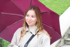 Laurence · 6960 x 4640 Cette photographie met en scène une jeune femme comme sujet principal, souriante et abritée sous un parapluie marron et rouge. La composition est simple, centrée sur son visage, avec un cadrage serré. L'arrière-plan flou montre un environnement extérieur verdoyant.La lumière semble naturelle, probablement en journée. Les couleurs dominantes sont les tons pastel de son vêtement, contrastant avec le rouge vif du parapluie. Le style est épuré et spontané, évoquant une ambiance détendue et joyeuse, malgré la présence du parapluie suggérant une légère pluie.