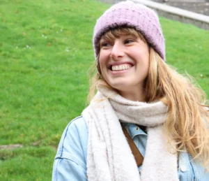 Laurence · 4876 x 4203 Cette photographie met en scène une jeune femme souriante, sujet principal, vêtue d’un pull bleu clair, d’un foulard blanc et d’un bonnet violet. Le cadrage est serré sur son visage et son haut, avec un arrière-plan flou de pelouse verte.La lumière semble naturelle et douce, éclairant son visage de manière homogène. Les couleurs dominantes sont les tons pastel (violet, bleu clair, blanc) et le vert de l’herbe. Le style est simple et spontané, évoquant une ambiance chaleureuse et détendue, presque printanière ou automnale.