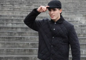 Laurence · 5790 x 4083 Cette photographie met en scène un jeune homme comme sujet principal, vêtu d’un style urbain et décontracté. Il porte un blouson noir matelassé et un béret, posant devant un mur en briques grises.Le cadrage est serré, centré sur lui, avec une légère inclinaison vers le haut. La lumière semble naturelle, douce et légèrement tamisée, créant des ombres discrètes. Les couleurs dominantes sont les tons noirs et gris, renforçant une ambiance sobre et moderne. L’ensemble dégage une impression de style minimaliste et contemporain, avec une touche d’élégance nonchalante.