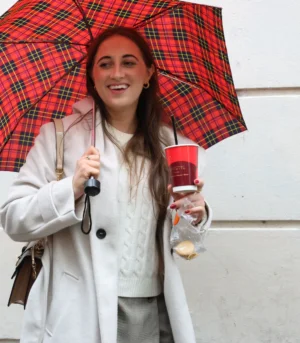 Laurence · 4058 x 4640 Cette photographie montre une jeune femme souriante, sujet principal, tenant un parapluie à carreaux rouges et noirs ainsi qu’un gobelet rouge. Elle porte un manteau clair et des gants.La composition est simple et centrée, avec un cadrage serré sur elle. La lumière semble naturelle, douce et légèrement diffuse, évitant les ombres marquées. Les couleurs dominantes sont les tons pastel (blanc, gris) et les couleurs vives (rouge, noir).L’ambiance est joyeuse et décontractée, évoquant une journée pluvieuse mais agréable. Le style est spontané et naturel.