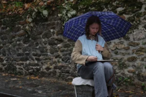 Laurence · 6960 x 4640 Cette photographie montre une personne assise sur un banc de pierre, protégée par un parapluie à carreaux bleus et blancs. Le sujet principal est cette personne, occupée à écrire dans un carnet.La composition est simple et naturelle, avec un cadrage serré qui isole le sujet. La lumière semble douce et diffuse, typique d’un jour nuageux. Les couleurs dominantes sont les tons naturels (vert des feuilles, gris de la pierre) et les bleus du parapluie.L’ambiance est calme et introspective, évoquant la sérénité et la concentration. Le style est réaliste et dépouillé.