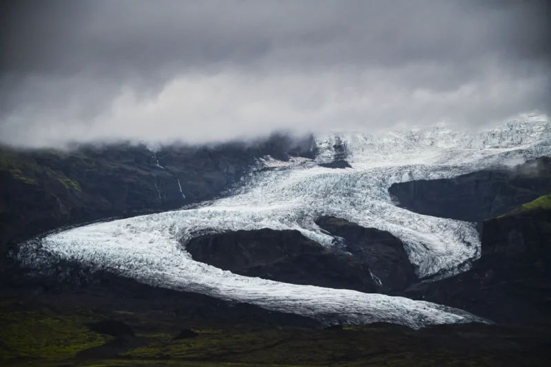 Ahmet · 29/08/2023 · 3000 x 2000 Cette photographie met en scène un glacier spectaculaire comme sujet principal. La composition montre une langue glaciaire sinueuse entourée de montagnes escarpées. Le cadrage est large, capturant une vue panoramique et immersive.La lumière est tamisée, avec des nuages bas créant une ambiance mystérieuse et dramatique. Les couleurs dominantes sont les teintes froides de blanc et de gris, accentuées par des nuances bleutées sur la glace. Le style est réaliste et naturel, évoquant une atmosphère sauvage et majestueuse, presque solennelle.