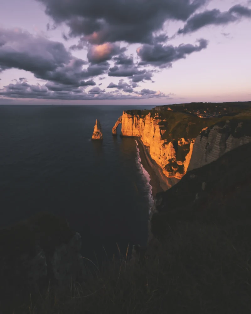 Ahmet · 23/09/2023 · 2400 x 3000 Cette photographie met en scène des **falaises côtières** éclairées par le soleil couchant. Le **sujet principal** est la lumière dorée qui illumine les parois rocheuses, contrastant avec l’obscurité de la mer et du ciel nuageux.La **composition** est équilibrée, avec un **cadrage serré** sur les falaises, accentuant leur verticalité. La **lumière rasante** crée des ombres profondes et des jeux de contrastes saisissants. Les **couleurs dominantes** sont les ors chauds, les gris bleutés et les noirs intenses.Le **style** est épuré et dramatique, évoquant une **ambiance mystique et puissante**, presque onirique.