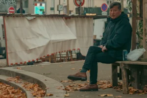 Don · 3853 x 2569 Cette photographie montre un homme assis seul sur un banc, sujet principal. La composition met en valeur son visage pensif et son attitude calme. Le cadrage est serré sur lui, avec un arrière-plan flou comprenant des éléments urbains et des feuilles mortes.La lumière semble naturelle, tamisée, créant une ambiance douce et légèrement mélancolique. Les couleurs dominantes sont des tons froids (bleu de son vêtement) et neutres (gris, beige), avec des touches de rouge et de blanc dans l’arrière-plan.Le style est réaliste et sobre, évoquant une ambiance contemplative et introspective.
