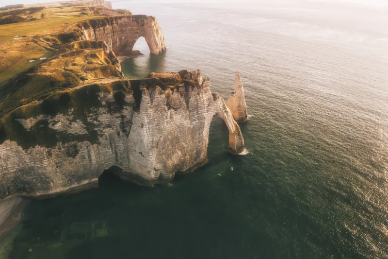 Ahmet · 27/06/2023 · 3000 x 1998 Cette photographie met en scène les **falaises d’Étretat**, avec comme sujet principal **l’arche naturelle** dominant la mer. La composition est dynamique, opposant la roche verticale et l’immensité océanique. Le cadrage aérien offre une vue plongeante, accentuant la grandeur du site.La lumière douce, probablement en fin de journée, adoucit les contours. Les couleurs dominantes sont les **tons ocres et verts** des falaises, contrastant avec le **bleu profond** de l’eau. Le style est épuré et naturel, évoquant une ambiance **sauvage et majestueuse**, presque mystique.