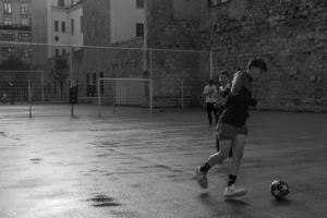 Don · 5072 x 3381 Cette photographie en noir et blanc capture une scène de football urbain. Le sujet principal est un joueur en action, contrôlant le ballon. La composition montre un terrain extérieur avec des bâtiments en arrière-plan, encadrés par des buts.Le cadrage est dynamique, mettant en valeur le mouvement du joueur. La lumière, naturelle et contrastée, accentue les ombres et les formes. Les couleurs dominantes, bien que monochromes, jouent sur les nuances de gris, renforçant l’ambiance dramatique et sportive. Le style est documentaire et spontané, évoquant une ambiance authentique et énergique de jeu improvisé.