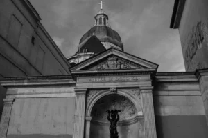 Don · 6918 x 4612 Cette photographie met en scène une façade architecturale sombre, probablement une église ou un bâtiment religieux. Le sujet principal est une entrée monumentale avec une arche et une statue centrale, encadrée par des colonnes.La composition est symétrique, avec un cadrage serré sur l’arche et la statue. La lumière, tamisée et contrastée, accentue les textures et les ombres, créant une ambiance dramatique. Les couleurs dominantes sont les tons de gris et noir, typiques d’un style photographique noir et blanc.L’ambiance est mystérieuse et solennelle, renforcée par les nuages menaçants dans le ciel.