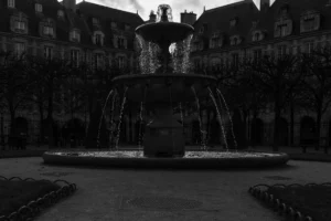 Don · 4750 x 3167 Cette photographie met en scène une **fontaine monumentale** comme sujet principal, avec une composition centrée et symétrique. Le cadrage est serré, encadrant la fontaine et ses jets d’eau.La lumière est faible, créant une **ambiance nocturne ou crépusculaire**, avec des contrastes marqués. Les **couleurs dominantes** sont les tons de gris et noir, typiques d’un cliché en noir et blanc.Le style est **sobre et dramatique**, renforcé par l’éclairage tamisé. L’ambiance évoque une certaine **mélancolie et sérénité**, accentuée par l’architecture urbaine en arrière-plan.