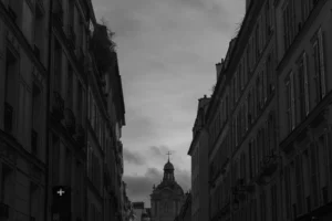 Don · 3352 x 2235 Cette photographie capture une rue étroite bordée de bâtiments anciens, formant une perspective convergente. Le sujet principal est une église ou un édifice religieux visible au loin, encadré par les immeubles sombres.Le cadrage est serré, créant un effet tunnel. La lumière est faible, avec des tons contrastés et une atmosphère dramatique. Les couleurs dominantes sont les gris et noirs, renforçant une ambiance mélancolique et mystérieuse. Le style rappelle le noir et blanc classique, évoquant une esthétique cinématographique ou documentaire. L’ambiance est à la fois sobre et poétique, mêlant solitude et grandeur architecturale.