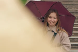 Don · 4948 x 3299 Cette photographie met en scène une jeune femme souriante, sujet principal, tenant un parapluie marron foncé. La composition est simple et centrée, avec un cadrage serré qui isole son visage et une partie de son haut.La lumière est douce et naturelle, probablement en extérieur par temps nuageux. Les couleurs dominantes sont les tons neutres (beige, marron) et les touches de vert et de rouge sur le parapluie. Le style est épuré et spontané, évoquant une ambiance chaleureuse et détendue.