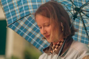 Don · 2557 x 1705 Cette photographie met en scène une jeune femme comme sujet principal, souriante et protégée par un parapluie à carreaux bleus et blancs. Le cadrage est serré sur son visage et le haut de son corps, avec un arrière-plan flou et légèrement verdâtre.La lumière semble naturelle, douce et légèrement tamisée, créant une ambiance chaleureuse. Les couleurs dominantes sont les tons pastel (bleu, blanc) du parapluie, contrastant avec les teintes naturelles de sa peau et ses cheveux roux. Le style est simple et spontané, évoquant une scène de vie quotidienne, pleine de douceur et de sérénité.