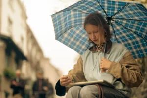 Don · 5909 x 3939 Cette photographie montre une femme comme sujet principal, assise sous un parapluie à carreaux bleus et blancs, lisant un livre ou un carnet. La composition est simple et centrée, avec un cadrage serré sur elle, légèrement flou en arrière-plan.La lumière semble naturelle, douce et tamisée, probablement par un temps nuageux. Les couleurs dominantes sont les tons froids (bleus, gris) et quelques touches chaudes (vêtements beiges et marron).Le style est réaliste et intimiste, évoquant une ambiance calme et contemplative, presque mélancolique.