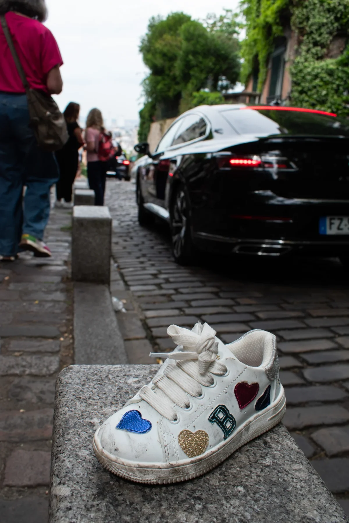 Deniz · 31/05/2025 · 1365 x 2048 Cette photographie met en avant une **chaussure blanche** décorée de motifs colorés (cœur, étoiles, drapeau) posée sur des pavés. Le **sujet principal** est ce sneaker, isolé au premier plan.La **composition** oppose ce détail intime à l’arrière-plan urbain animé : voitures, trottoir et passants. Le **cadrage** est serré sur la chaussure, avec une profondeur de champ qui floute légèrement l’arrière-plan.Les **couleurs dominantes** sont les tons neutres (gris des pavés, noir des voitures) contrastant avec les **couleurs vives** de la chaussure. La **lumière** semble naturelle, avec des ombres douces. L’ambiance est **urbaine et contrastée**, mêlant intimité et vie collective.