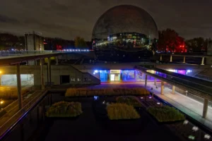 Cette photographie capture une scène nocturne urbaine avec pour sujet principal un grand dôme architectural lumineux en arrière-plan, probablement le Millennium Dome à Londres.La composition montre une perspective depuis un pont ou une passerelle, avec des bâtiments éclairés et des néons colorés en bas. Le cadrage inclut des éléments urbains comme des voitures et des structures modernes. Les lumières, vives et contrastées, créent une ambiance dynamique. Les couleurs dominantes sont les bleus, rouges et blancs, renforçant l’aspect futuriste et vibrant. Le style est photographique urbain nocturne, évoquant une ambiance énergique et moderne.