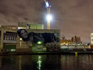 Cette photographie montre un bâtiment industriel la nuit avec une projection géante d’un visage féminin sur sa façade. Le sujet principal est cette projection lumineuse contrastant avec l’architecture sombre.La composition joue sur le cadrage serré du bâtiment et de l’eau en premier plan. Les lumières artificielles, notamment le faisceau du projecteur et les éclairages urbains, dominent, créant une ambiance mystérieuse et moderne. Les couleurs sont principalement sombres (noirs, gris) avec des éclats de bleu et de blanc, renforçant l’atmosphère nocturne et industrielle. Le style est réaliste et contemporain, évoquant une touche artistique et urbaine.
