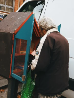 Ahmet · 22/11/2025 · 1919 x 2560 Cette photographie montre une personne âgée, sujet principal, en train d’interagir avec une petite structure colorée en bois, probablement un distributeur ou une boîte à lettres originale. La composition est simple, avec un cadrage serré sur le sujet et l’objet.La lumière semble naturelle, un peu diffuse, créant des contrastes doux. Les couleurs dominantes sont les teintes vives du bois (bleu, rouge) et les tons neutres du vêtement de la personne. L’ambiance est chaleureuse et quotidienne, évoquant une scène de rue paisible et authentique. Le style est documentaire et spontané.