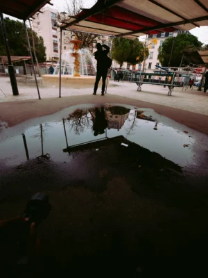 Ahmet · 22/11/2025 · 1919 x 2560 Cette photographie capture une flaque d’eau au premier plan reflétant partiellement un environnement urbain. Le sujet principal est cette surface liquide, agissant comme un miroir déformant.La composition joue sur la symétrie et la distorsion, avec un cadrage en contre-plongée. L’arrière-plan montre des personnes sous une structure couverte, avec des couleurs dominées par des tons neutres (gris, beige) et des éclats de rouge et blanc.La lumière semble naturelle, tamisée, créant une ambiance calme et légèrement mélancolique. Le style est réaliste et documentaire, évoquant une scène du quotidien.