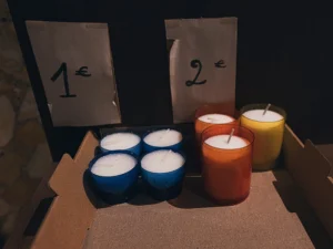Ahmet · 22/11/2025 · 2560 x 1919 Cette photographie montre principalement des bougies artisanales disposées dans des cartons. Le sujet inclut quatre bougies teintées (bleues et jaunes) et deux bougies plus hautes (orange et jaune).La composition est simple et épurée, avec un cadrage serré sur les bougies et leurs étiquettes de prix (1 € et 2 €). La lumière est tamisée, créant une ambiance chaleureuse et intimiste. Les couleurs dominantes sont les teintes vives des bougies, contrastant avec le fond sombre. Le style est minimaliste et artisanal, évoquant une ambiance artisanale et conviviale.