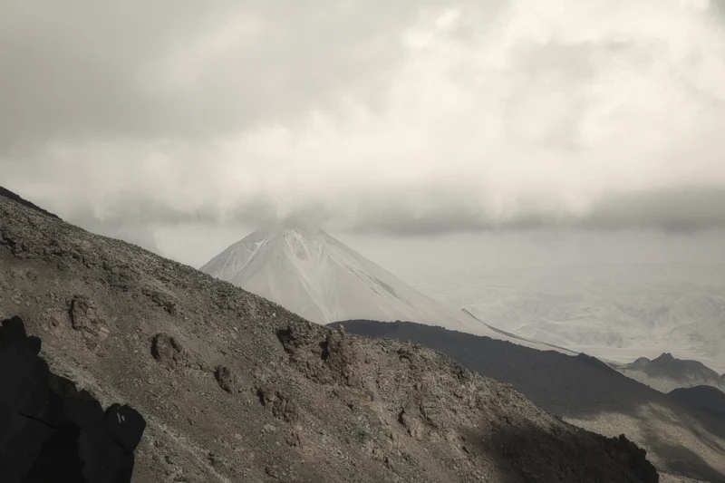 Ahmet · 14/08/2025 · 3000 x 1999 Cette photographie met en scène un **volcan enneigé** comme sujet principal, partiellement émergent des nuages. La **composition** joue sur les contrastes entre la roche sombre en premier plan et la neige/le ciel en arrière-plan.Le **cadrage** est serré sur la base du volcan, créant une perspective dramatique. La **lumière**, douce et diffuse, adoucit les contours, tandis que les **couleurs dominantes** sont les gris, blancs et noirs, renforçant une ambiance mystérieuse et minérale.Le style est **sobre et naturel**, évoquant une atmosphère **sauvage et majestueuse**, presque onirique.