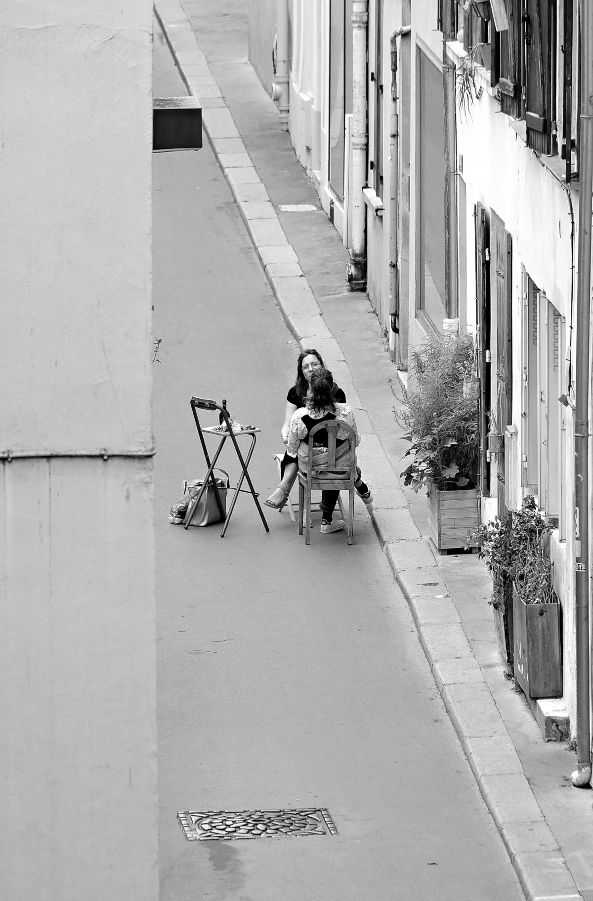 AnJo · 09/06/2022 · 2630 x 4000 Cette photographie en noir et blanc met en scène une personne assise sur un vélo, poussée par un enfant. Le sujet principal est cette scène de rue paisible.La composition est simple et équilibrée, avec un cadrage serré sur le côté de la rue. La lumière naturelle, douce, crée des contrastes modérés. Les couleurs dominantes, bien que monochromes, varient du noir au gris clair.Le style est documentaire et intemporel, évoquant une ambiance calme et quotidienne, presque nostalgique. L'image capte une scène authentique et intimiste.