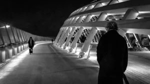Cette photographie en noir et blanc met en scène deux silhouettes sur un pont moderne et lumineux. Le sujet principal semble être la personne dos à la caméra, soulignant une impression de solitude ou de contemplation. La composition joue sur les lignes architecturales du pont, créant une perspective dynamique. Le cadrage serré et les jeux d'ombres accentuent le contraste entre lumière et obscurité. Les couleurs dominantes, bien que monochromes, exploitent les nuances de gris pour renforcer l’ambiance nocturne et mystérieuse. Le style est épuré et cinématographique, évoquant une atmosphère mélancolique et introspective.