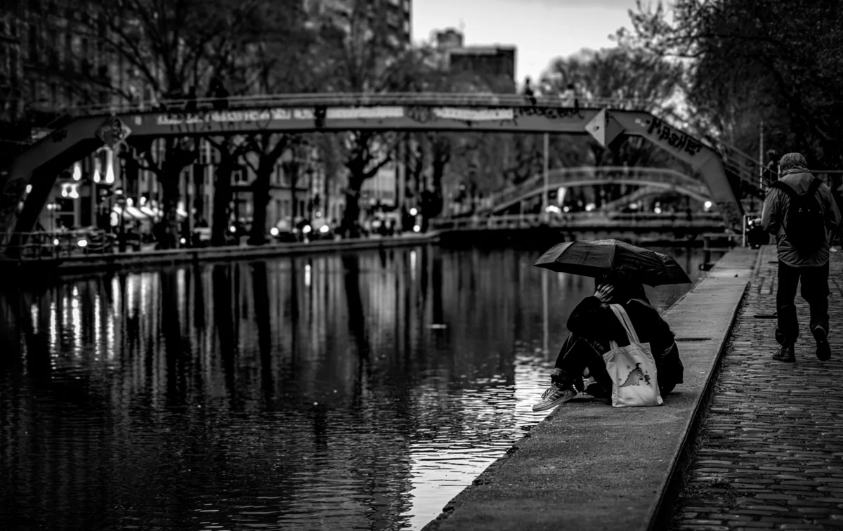 AnJo · 29/03/2024 · 5508 x 3467 Cette photographie en noir et blanc met en scène un **sujet principal** : une personne penchée sur le bord d’un canal, probablement pour nourrir des canards. La **composition** est équilibrée, avec le chemin en pierre menant vers un **pont** à l’arrière-plan. Le **cadrage** est serré sur le sujet et son environnement immédiat, créant une ambiance intime.La **lumière**, douce et tamisée, suggère un moment crépusculaire ou nocturne. Les **couleurs dominantes** sont les nuances de gris, renforçant une atmosphère mélancolique et paisible. Le **style** est documentaire et poétique, évoquant une scène du quotidien. L’**ambiance** est calme et contemplative, presque nostalgique.