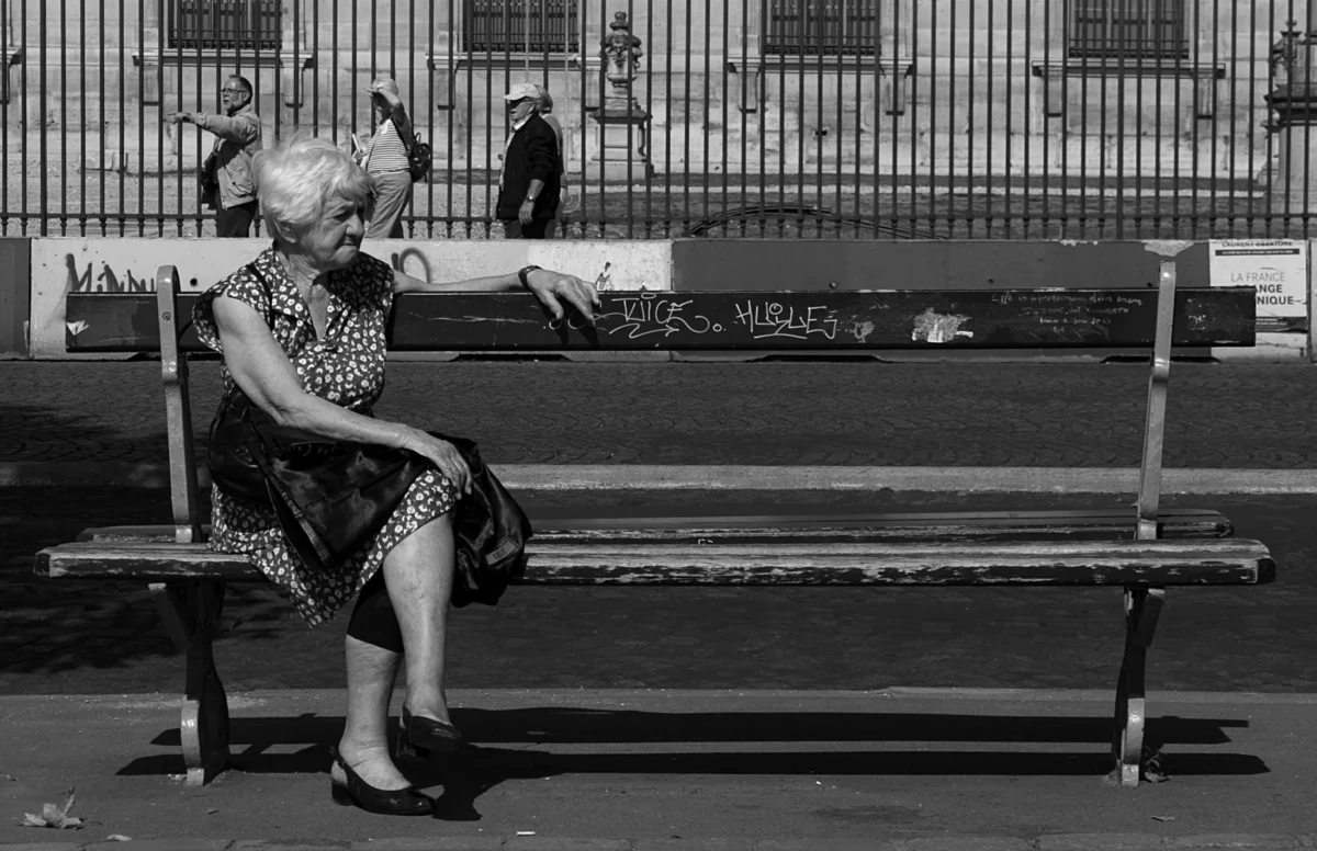 AnJo · 11/09/2015 · 3311 x 2141 Cette photographie en noir et blanc met en scène une personne âgée, sujet principal, assise sur un banc public. Elle semble absorbée par un objet entre ses mains.La composition est simple et centrée, avec un cadrage serré sur le sujet et le banc. L’arrière-plan, légèrement flou, montre des graffitis et une barrière métallique.La lumière est naturelle, contrastée, mettant en valeur les textures et les ombres. Les couleurs dominantes, bien que monochromes, jouent sur les nuances de gris.Le style est documentaire et intime, évoquant une ambiance mélancolique et contemplative.