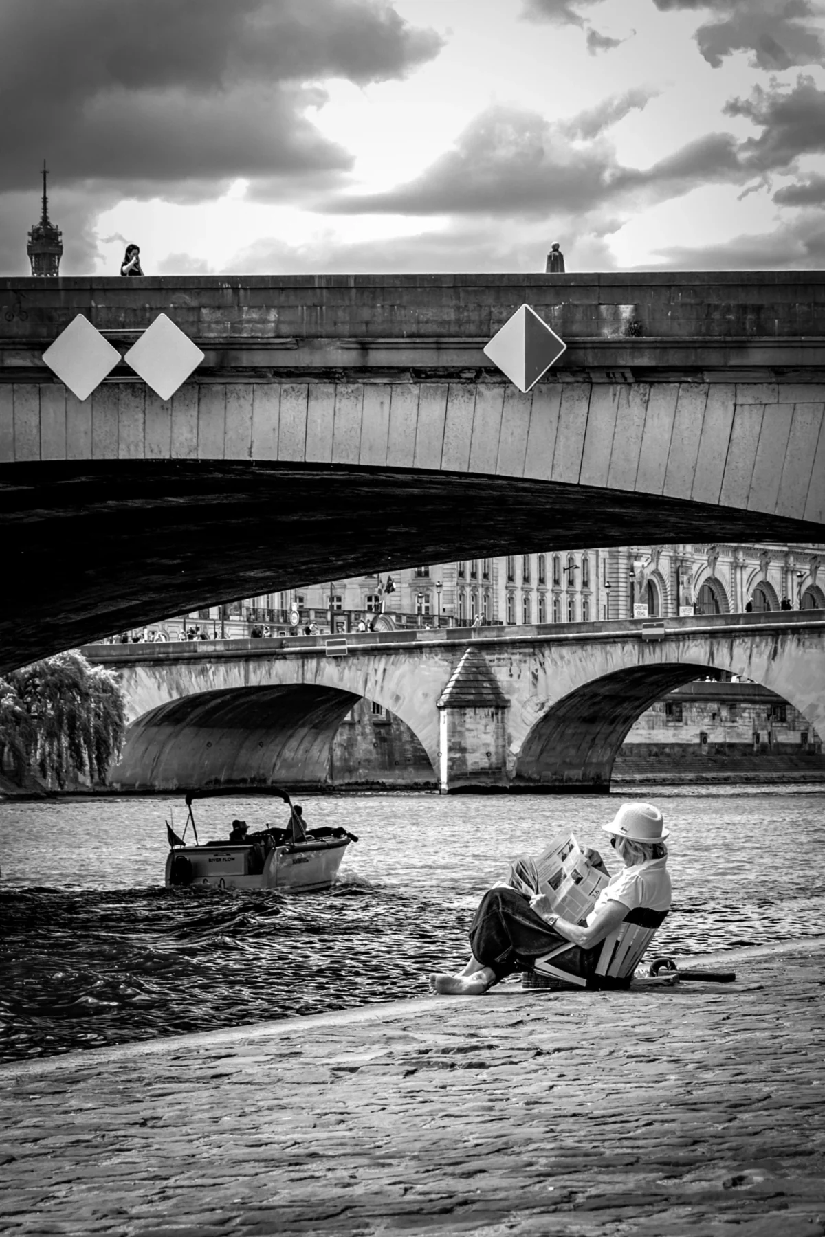 AnJo · 11/05/2025 · 3474 x 5211 Cette photographie en noir et blanc met en scène un homme assis sur une péniche ou un bateau sur un fleuve, sous un pont. Le sujet principal est cet individu, absorbé dans une activité calme, probablement la pêche.La composition joue sur les lignes horizontales du pont et de l’eau, créant une perspective profonde. Le cadrage inclut aussi un autre bateau à gauche, ajoutant de la dynamique.La lumière est douce, avec des contrastes marqués entre ombres et clartés, renforçant l’ambiance contemplative. Les tons dominants sont les gris et noirs, typiques du noir et blanc, accentuant le côté intemporel et mélancolique. Le style est documentaire et poétique, évoquant une atmosphère paisible et nostalgique.