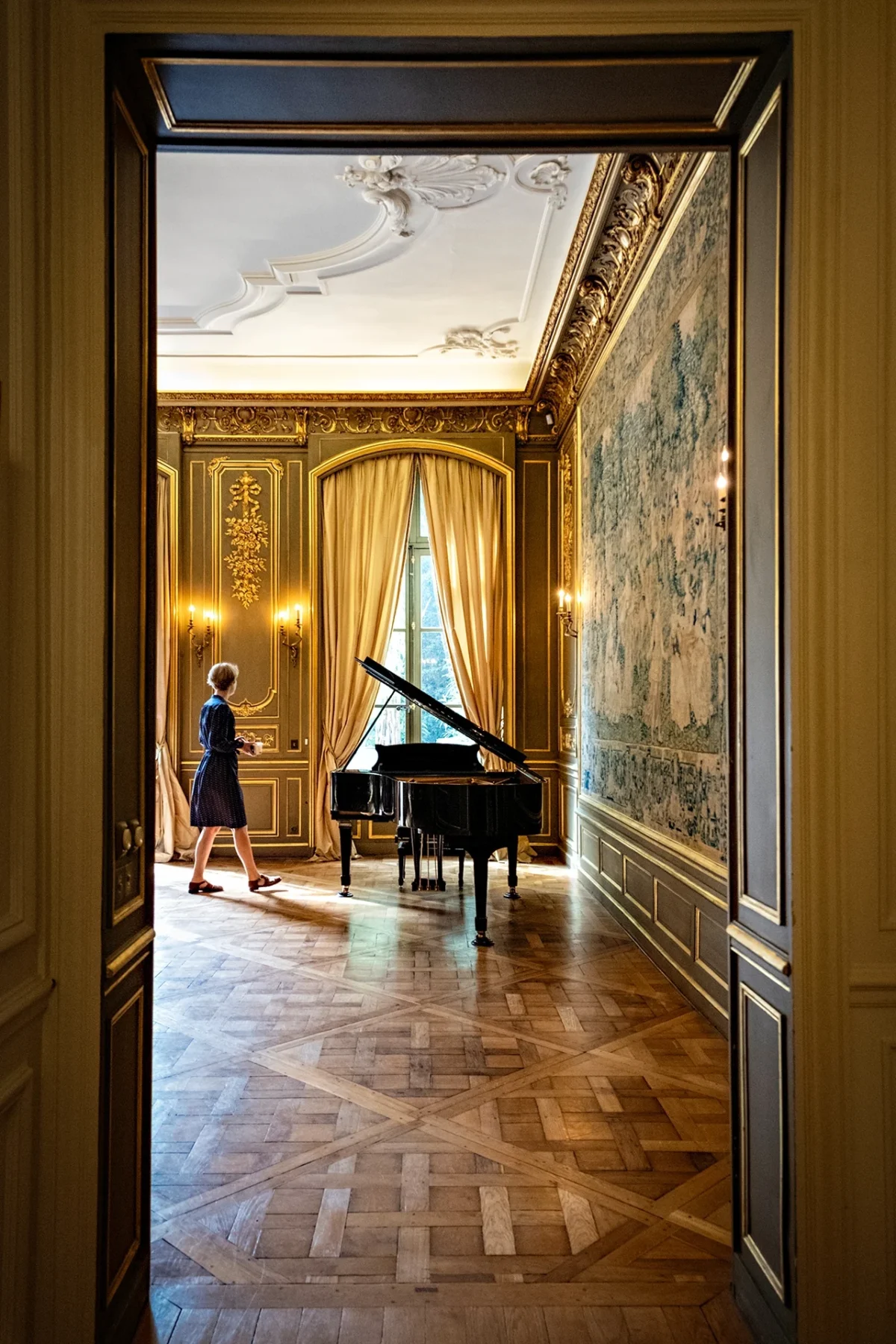 AnJo · 21/09/2024 · 1280 x 1920 Cette photographie met en scène une femme jouant du piano dans un salon élégant. Le sujet principal est le piano et la musicienne, située en profondeur.La composition utilise un cadrage en profondeur avec une perspective convergente, accentuée par le plancher en damier. La lumière, douce et naturelle, provient probablement d’une source externe, éclairant la scène de manière chaleureuse. Les couleurs dominantes sont des tons neutres (beige, gris) rehaussés par des dorures et des touches de bleu-vert sur les murs.Le style est classique et raffiné, évoquant une ambiance luxueuse et intemporelle, presque théâtrale.