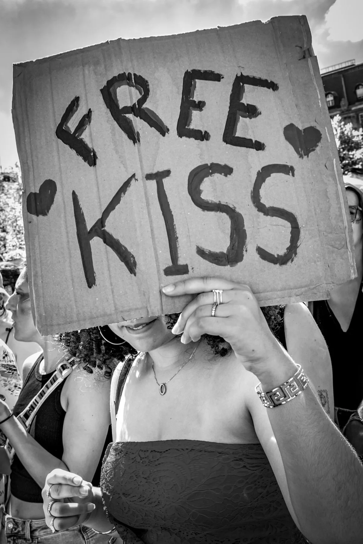 AnJo · 28/06/2025 · 2666 x 4000 Cette photographie montre une personne tenant un panneau avec écrit **"FREE KISS"** en lettres manuscrites, entourées de cœurs. Le sujet principal est flou, mettant l’accent sur le message.La composition est centrée sur le panneau, avec un cadrage serré. La lumière est naturelle, contrastée, créant des ombres marquées. En noir et blanc, les tons varient du clair au foncé, accentuant les détails et les textures.Le style est documentaire et spontané, évoquant une ambiance militante ou festive, avec une touche d’ironie et de liberté.
