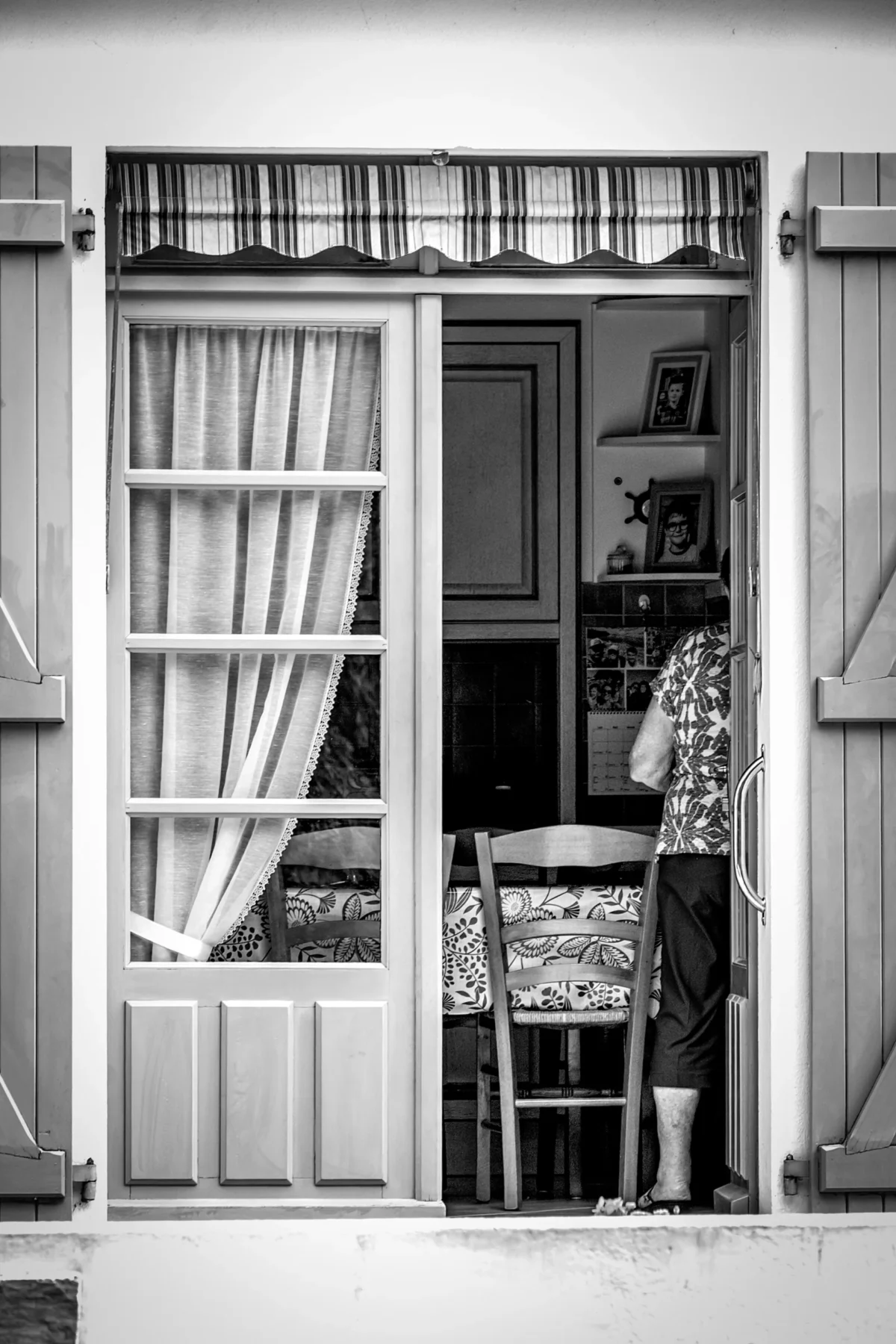 AnJo · 02/08/2025 · 2667 x 4000 Cette photographie en noir et blanc met en scène un intérieur vu à travers une fenêtre ouverte. Le sujet principal est une chaise en osier et un petit espace intérieur avec des éléments décoratifs, comme une table et des objets divers.La composition est symétrique et équilibrée, avec un cadrage serré sur la fenêtre. La lumière naturelle, douce et tamisée, traverse les rideaux, créant des contrastes subtils. Les couleurs dominantes, bien que monochromes, jouent sur les nuances de gris et de blanc.Le style est documentaire et intemporel, évoquant une ambiance calme et nostalgique, presque poétique.