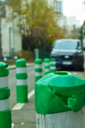Deniz · 22/11/2025 · 3838 x 5757 Cette photographie met en scène un **bollard vert** recouvert d’un **sac vert transparent** déchiré, posant sur un trottoir. Le **sujet principal** est ce sac, symbolisant peut-être un problème environnemental.La **composition** est simple, avec un **cadrage serré** sur le premier plan. En arrière-plan, on aperçoit une **rue avec des bollards alignés**, une voiture et des bâtiments flous. La **lumière** semble naturelle, légèrement tamisée, créant des ombres douces.Les **couleurs dominantes** sont le **vert vif** (bollards et sac) et des tons neutres (gris du trottoir, blanc des bâtiments). Le style est **réaliste et documentaire**, évoquant une **ambiance urbaine et légèrement mélancolique**.