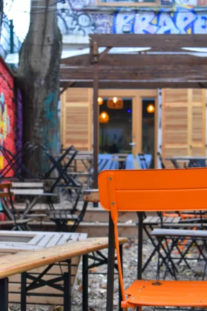 Deniz · 22/11/2025 · 4000 x 6000 Cette photographie met en scène un **café ou une terrasse urbaine**. Le **sujet principal** est une table en bois avec des chaises orange, partiellement à l’ombre. La **composition** montre un arrière-plan flou avec des graffitis et des structures en bois, évoquant un lieu artistique ou alternatif.Le **cadrage** est serré, centré sur la table, avec une profondeur de champ réduite. La **lumière** est douce et naturelle, avec des ombres marquées. Les **couleurs dominantes** sont les tons bois, orange vif, et des touches de bleu et vert des graffitis.L’ambiance est **urbaine, décontractée et artistique**, mêlant intimité et créativité.