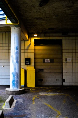 Deniz · 22/11/2025 · 3856 x 5784 Cette photographie montre un espace souterrain ou semi-enterré, probablement un parking ou un local technique. Le sujet principal est une porte métallique entrouverte, éclairée par une lumière artificielle jaunâtre.La composition est simple et symétrique, avec des murs carrelés de couleur crème et des éléments industriels comme une colonne peinte en bleu et jaune. Le cadrage est serré, mettant en valeur les détails architecturaux et les ombres marquées. Les couleurs dominantes sont les tons froids (gris, bleu) et chauds (jaune, blanc). L’ambiance est austère et fonctionnelle, évoquant un lieu peu fréquenté et utilitaire.